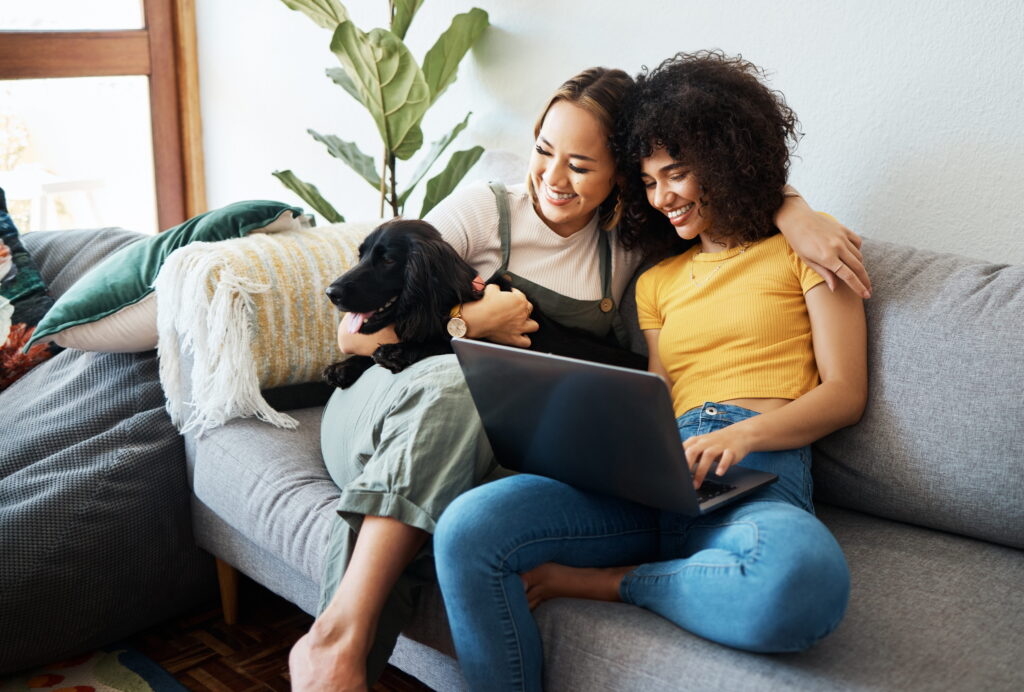 Two women sitting on a couch with a black dog looking happily at a laptop