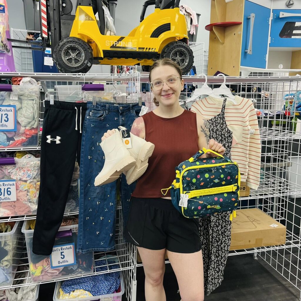 Woman holding a lunchbox and shoes standing in front of a back-to-school display at Once Upon a Child in Coralville, Iowa